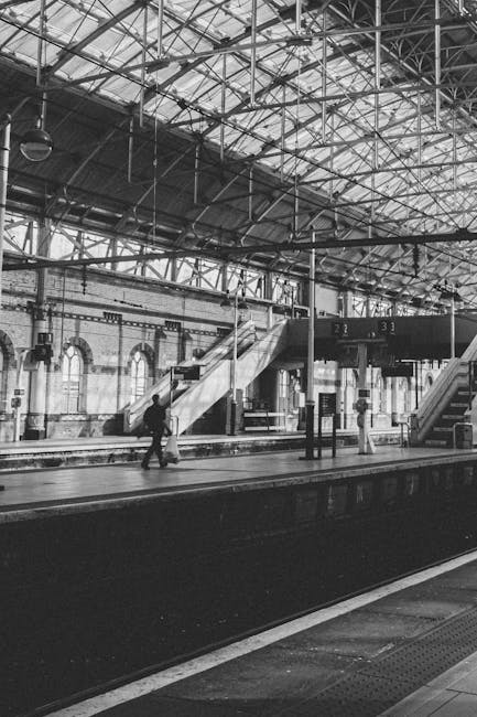 A black-and-white photograph of Anerley station, showing the interior with a high, arched metal roof structure supported by beams and girders. The station platform is visible with a few passengers, including one man walking with a small wheeled trolley, and another standing nearby. The platform features a staircase leading to an upper level and additional stairs ascending to the left, with platform signage displaying platform numbers 2 and 3. Adjacent to the platform, a row of large windows lets in natural light, illuminating the space. Tracks run parallel to the platform in the foreground, and the overall environment is typical of an urban train station used for home relocation tasks, furniture transport, and luggage movement. The scene captures the logistical process of loading and unloading belongings in a tenant or homeowner moving context, supported by Man with Van Crystal Palace as a professional removals service.