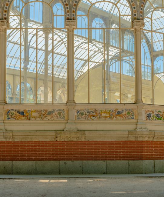 A detailed exterior view of a large, historic greenhouse or conservatory with ornate metal framing and extensive glass panels, set against a bright blue sky. The structure features arched windows and 
