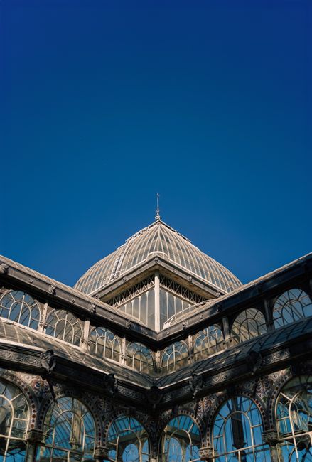 The image shows a close-up view of a historic glass and metal conservatory or greenhouse structure with ornate arched windows and decorative metalwork, situated at the top of a building with a promine