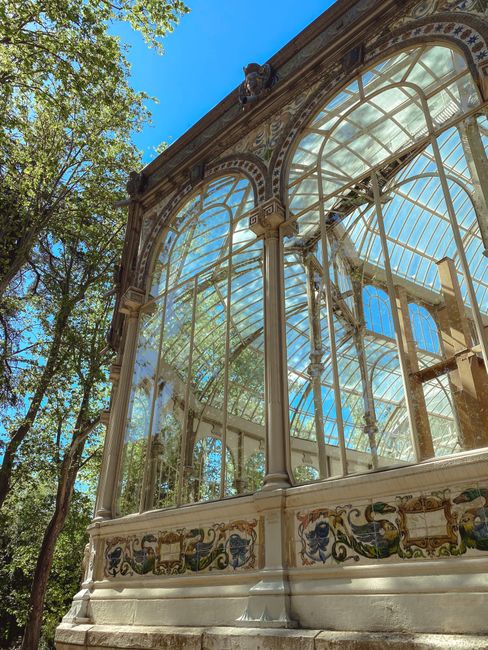 A detailed view of an ornate, historic glass conservatory with a curved, arched roof made of transparent glass panels and decorative metal framework. The structure features tall windows with multiple