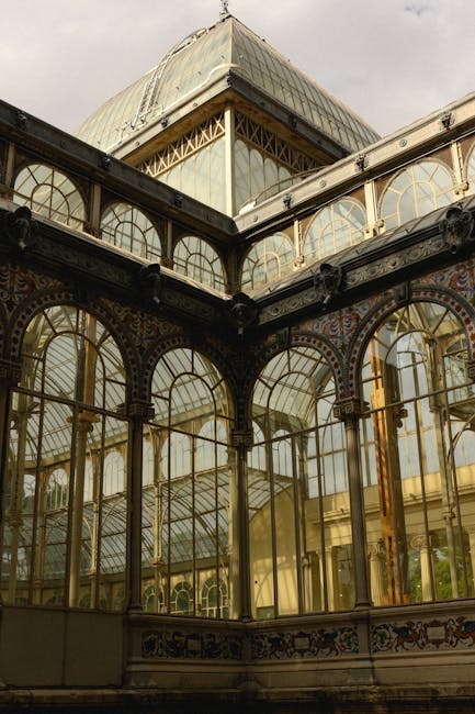 A detailed view of an ornate, historic glass conservatory with a curved, arched roof made of transparent glass panels and decorative metal framework. The structure features tall windows with multiple